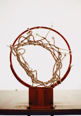 A close-up view of a basketball hoop, focusing on the rim and the net. The net appears old and frayed, with some strings hanging loose. The background is plain and light, allowing the hoop and net to stand out prominently.
