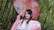 A person wearing a traditional outfit stands amidst lush green bamboo plants. They hold an ornate pink and white parasol with designs above their head. Their attire is adorned with floral embroidery, and they have decorative hair accessories.