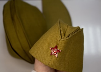 A Soviet military-style cap is adorned with a red star pin featuring a hammer and sickle emblem. The cap is olive green and appears to be made of a textured fabric. Another similar cap is visible in the background.