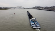 A large cargo ship is navigating a wide river, carrying a load of coal. In the background, a bridge spans across the river, with a cityscape lining the right bank. The sky is overcast, hinting at a gloomy atmosphere.