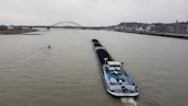 A large cargo ship is navigating a wide river, carrying a load of coal. In the background, a bridge spans across the river, with a cityscape lining the right bank. The sky is overcast, hinting at a gloomy atmosphere.