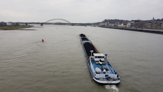 A large cargo ship is navigating a wide river, carrying a load of coal. In the background, a bridge spans across the river, with a cityscape lining the right bank. The sky is overcast, hinting at a gloomy atmosphere.