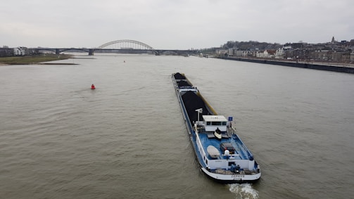 A large cargo ship is navigating a wide river, carrying a load of coal. In the background, a bridge spans across the river, with a cityscape lining the right bank. The sky is overcast, hinting at a gloomy atmosphere.
