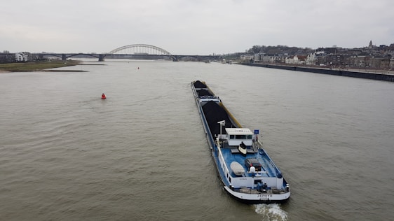 A large cargo ship is navigating a wide river, carrying a load of coal. In the background, a bridge spans across the river, with a cityscape lining the right bank. The sky is overcast, hinting at a gloomy atmosphere.