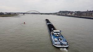 A large cargo ship is navigating a wide river, carrying a load of coal. In the background, a bridge spans across the river, with a cityscape lining the right bank. The sky is overcast, hinting at a gloomy atmosphere.