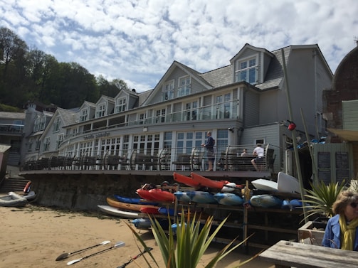 A large, three-story building with multiple windows and a deck overlooking a sandy area. Several colorful kayaks are stacked near the deck, and a few people are visible enjoying the outdoor space. Tall trees can be seen in the background under a partly cloudy sky.