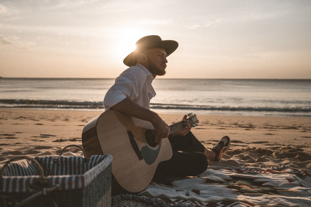 uomo in camicia bianca e cappello da cowboy marrone che suona la chitarra acustica sulla spiaggia durante il giorno