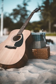 An acoustic guitar rests against a woven basket on a sandy beach, with blurred foliage in the background under a clear sky.