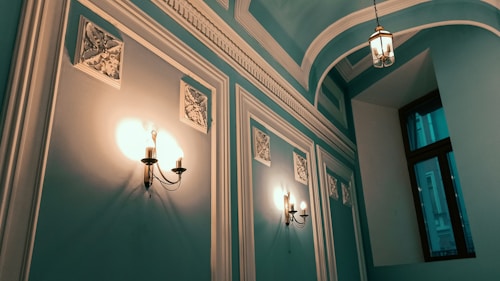 An interior hallway with teal-colored walls featuring white ornate molding and decorative panels. The walls are adorned with vintage wall-mounted lamps emitting a warm yellow light. The ceiling is arched with intricate molding, and a single hanging light fixture is visible. A large window with a black frame is on the right side of the hallway.