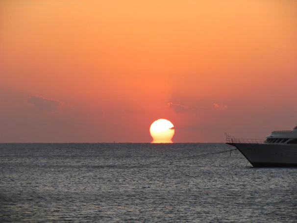 Sunset over an Aegean cove with a yacht silhouetted against orange and pink sky reflections on the water.