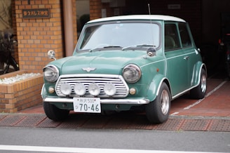 A vintage green Mini Cooper car is parked on a brick-paved driveway in front of a building with a brick facade. The car has a distinct classic design with round headlights and a noticeable front grille featuring multiple mounted spotlights. The license plate is prominently displayed with Japanese characters and numbers. To the left, there appears to be a small garden area with white stones and a bicycle partially visible.