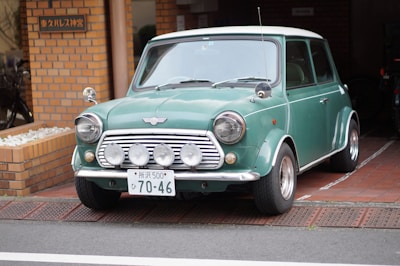 A vintage green Mini Cooper car is parked on a brick-paved driveway in front of a building with a brick facade. The car has a distinct classic design with round headlights and a noticeable front grille featuring multiple mounted spotlights. The license plate is prominently displayed with Japanese characters and numbers. To the left, there appears to be a small garden area with white stones and a bicycle partially visible.