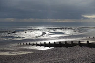 A serene and expansive beach scene with a pebbled shoreline leading to a calm sea. Wooden groynes extend towards the ocean, and the surface of the water reflects the overcast sky above. Dark clouds loom but hints of light break through, casting a silvery sheen on the wet sand.