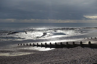 A serene and expansive beach scene with a pebbled shoreline leading to a calm sea. Wooden groynes extend towards the ocean, and the surface of the water reflects the overcast sky above. Dark clouds loom but hints of light break through, casting a silvery sheen on the wet sand.