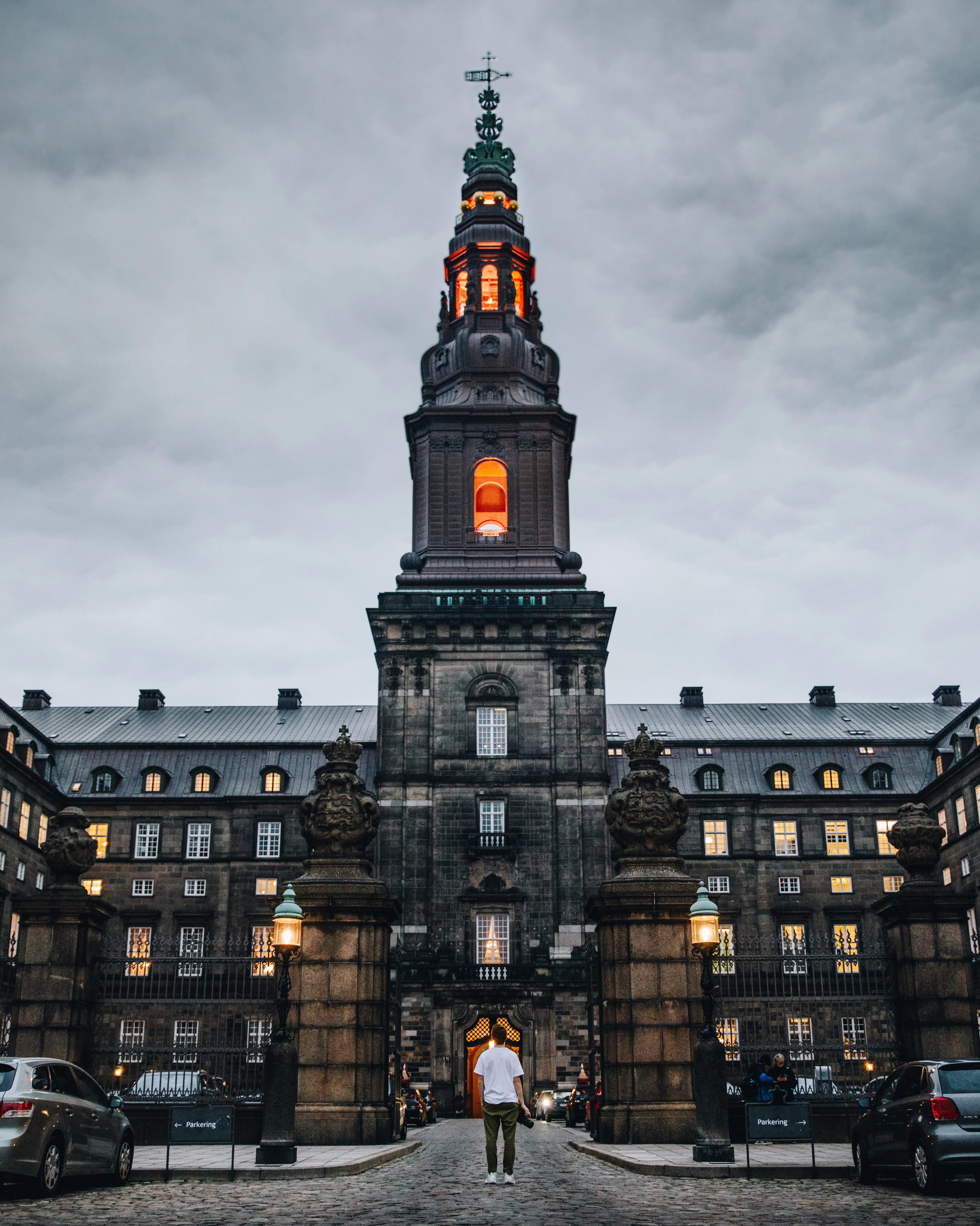 A grand spired palace glows at dusk, windows lit warm orange, a lone figure walking toward its gate.