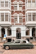 A vintage car is parked on a cobblestone street in front of a historic building with ornate windows and decorative brickwork. The building is multi-storied and features white window frames and intricate architectural details. A bicycle is leaning against the wall near the entrance.