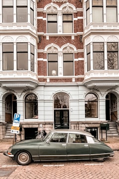 A vintage car is parked on a cobblestone street in front of a historic building with ornate windows and decorative brickwork. The building is multi-storied and features white window frames and intricate architectural details. A bicycle is leaning against the wall near the entrance.