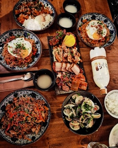 A wooden table is lavishly set with a variety of Korean dishes. There are several bowls containing rice, seasoned meat, noodles topped with eggs, and clams, all garnished with herbs and vegetables. A platter of grilled meats and kimchi is also present, along with bowls of a pale beverage. A bottle of traditional Korean drink, possibly rice wine, is also visible.