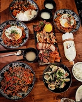 A wooden table is lavishly set with a variety of Korean dishes. There are several bowls containing rice, seasoned meat, noodles topped with eggs, and clams, all garnished with herbs and vegetables. A platter of grilled meats and kimchi is also present, along with bowls of a pale beverage. A bottle of traditional Korean drink, possibly rice wine, is also visible.