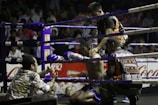 A boxing ring scene with a fighter sitting on a stool in the corner, wearing purple gloves and shorts. Three other individuals surround him, appearing to provide support and encouragement. The setting is a crowded arena with spectators visible in the background.