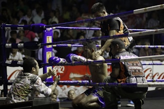 A boxing ring scene with a fighter sitting on a stool in the corner, wearing purple gloves and shorts. Three other individuals surround him, appearing to provide support and encouragement. The setting is a crowded arena with spectators visible in the background.
