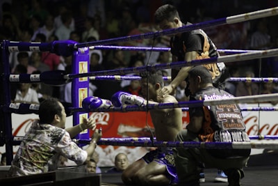 A boxing ring scene with a fighter sitting on a stool in the corner, wearing purple gloves and shorts. Three other individuals surround him, appearing to provide support and encouragement. The setting is a crowded arena with spectators visible in the background.