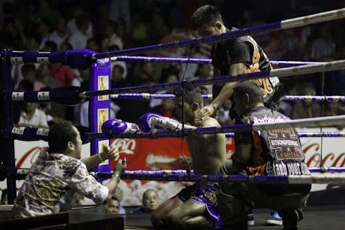 A boxing ring scene with a fighter sitting on a stool in the corner, wearing purple gloves and shorts. Three other individuals surround him, appearing to provide support and encouragement. The setting is a crowded arena with spectators visible in the background.