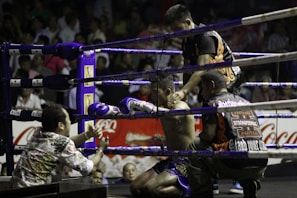 A boxing ring scene with a fighter sitting on a stool in the corner, wearing purple gloves and shorts. Three other individuals surround him, appearing to provide support and encouragement. The setting is a crowded arena with spectators visible in the background.