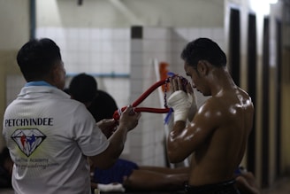 A shirtless man with wrapped hands is holding a traditional headpiece, suggesting a ritual or moment of focus before a fight. Another person stands nearby, wearing a shirt with a logo, likely indicating association with a martial arts academy. The setting appears to be a dimly lit gym or training area.