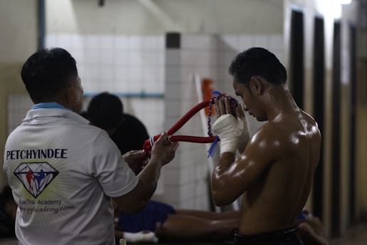 A shirtless man with wrapped hands is holding a traditional headpiece, suggesting a ritual or moment of focus before a fight. Another person stands nearby, wearing a shirt with a logo, likely indicating association with a martial arts academy. The setting appears to be a dimly lit gym or training area.