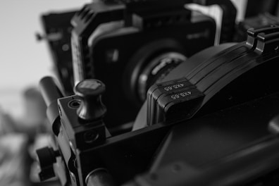 A close-up view of a professional camera setup, focusing on the lens and attached equipment. The image is in black and white, highlighting the intricate details of the metal and plastic components. The numbers 4x5.65 are visible on a part of the equipment.