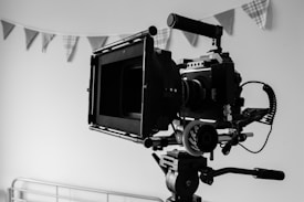 A professional video camera setup on a tripod inside a room. The camera is equipped with various attachments and faces the center of the room, suggesting it is ready for filming. Behind the camera, a string of decorative triangular flags hang against the wall, adding a touch of festivity to the otherwise minimal setting.