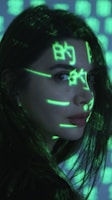 A person is seen with bright green digital symbols projected onto their face and hair. The symbols resemble characters from a computer screen, giving a futuristic or cybernetic appearance against the dimly lit background. The person has long dark hair and is looking sideways.