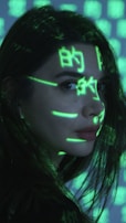 A person is seen with bright green digital symbols projected onto their face and hair. The symbols resemble characters from a computer screen, giving a futuristic or cybernetic appearance against the dimly lit background. The person has long dark hair and is looking sideways.