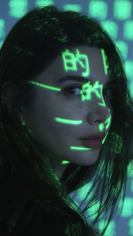 A person is seen with bright green digital symbols projected onto their face and hair. The symbols resemble characters from a computer screen, giving a futuristic or cybernetic appearance against the dimly lit background. The person has long dark hair and is looking sideways.