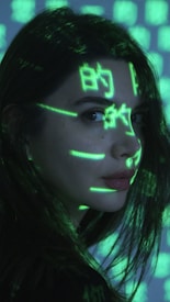 A person is seen with bright green digital symbols projected onto their face and hair. The symbols resemble characters from a computer screen, giving a futuristic or cybernetic appearance against the dimly lit background. The person has long dark hair and is looking sideways.