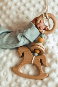 A baby's hand is holding a wooden toy featuring a ring and a rocking horse shape, adorned with colorful beads. The toy rests on a soft, textured white surface, and the baby is wearing a light-colored, ribbed fabric top.