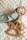 A baby's hand is holding a wooden toy featuring a ring and a rocking horse shape, adorned with colorful beads. The toy rests on a soft, textured white surface, and the baby is wearing a light-colored, ribbed fabric top.