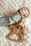 A baby's hand is holding a wooden toy featuring a ring and a rocking horse shape, adorned with colorful beads. The toy rests on a soft, textured white surface, and the baby is wearing a light-colored, ribbed fabric top.