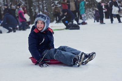 girl in black jacket and blue denim jeans sitting on white snow during daytime sled zoom background