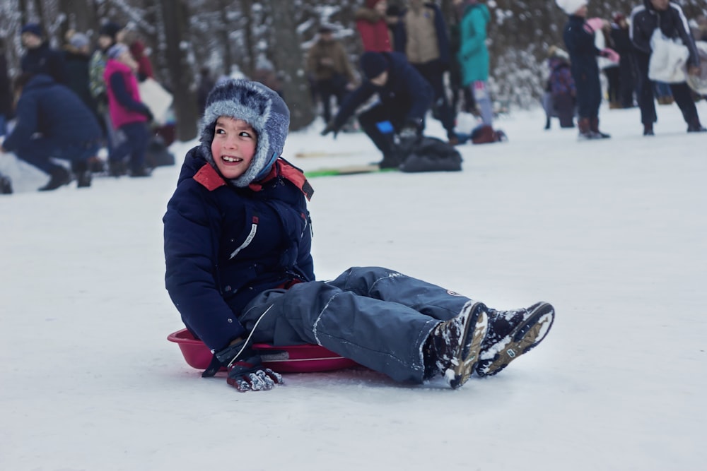 Mädchen in schwarzer Jacke und blauen Jeans sitzt tagsüber auf weißem Schnee