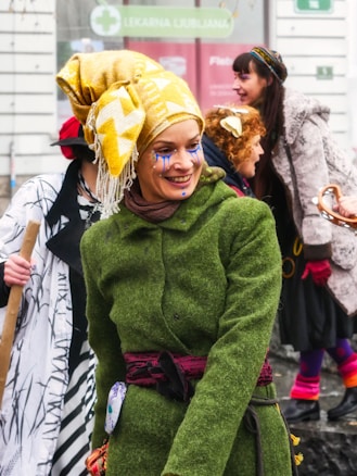 A group of people dressed in colorful, eclectic costumes. The central figure wears a bright yellow headwrap, green textured coat, and has blue face paint. Others in the background also have vibrant attires and are engaged in joyful interaction.