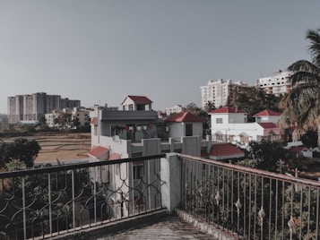 A scenic view from a balcony overlooking residential houses with red roofs, surrounded by greenery and palm trees. High-rise apartment buildings are visible in the background under a clear sky, while a field with crops lies adjacent to the houses.