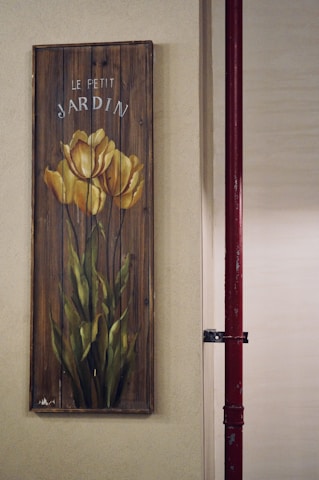 A vertical wooden panel displays a painted image of yellow tulips with green stems and leaves. The words 'Le Petit Jardin' are inscribed above the flowers. The panel appears to be attached to a textured beige wall. To the right, part of a red metal pipe is visible.