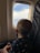 A child looks out of an airplane window. The child is wearing a face mask and has a shirt with anchor designs. Through the window, a clear blue sky with some clouds is visible. The child holds a colorful blanket.