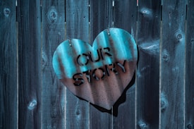 A heart-shaped metal sign with cut-out letters spelling 'OUR STORY' is mounted on a dark wooden fence. The metal has a blue-green patina, contributing to a rustic aesthetic.