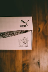 A cardboard box with a grid pattern on the exterior features the logo of a well-known sportswear brand. There is a black and white sketch on the box depicting two cartoon-like creatures. The background is a wooden surface.