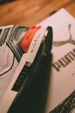A close-up of a sneaker with detailed mesh and fabric patterns in shades of white, gray, and black, featuring an orange detail with branding and an artist's signature on the side. The sneaker is placed on top of a box with a visible logo.