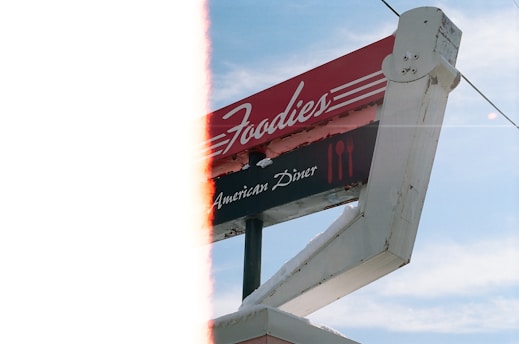 A slightly damaged vintage sign for an American diner named 'Foodies' is displayed against a clear blue sky. The sign features red and white colors with icons of a fork and knife. Part of the image has a light leak effect.