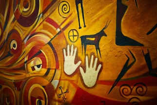 A vibrant cave painting that includes handprints, abstract swirl patterns, geometric symbols, and a depiction of an animal, possibly a deer. The artwork is set against a predominantly warm background, with shades of yellow, orange, and red.
